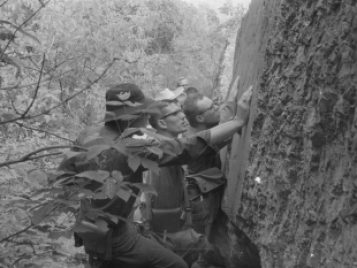 A black-and-white photograph of young men inspecting a large rock. They all appear to be looking at the same feature.